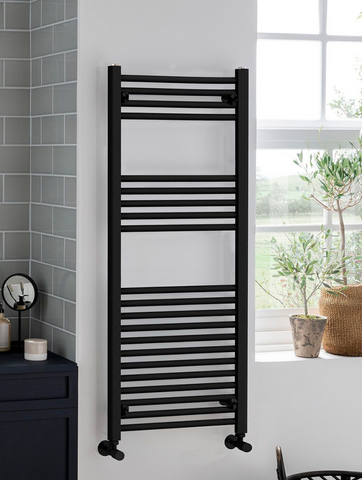 Black towel radiator mounted on a white wall next to a window with plants.
